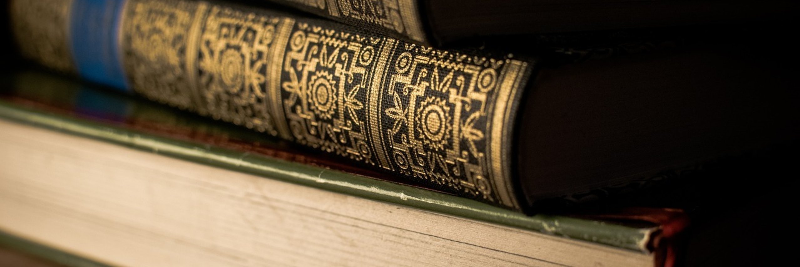 In Nahaufnahme sind drei übereinanderliegende, historische Bücher zu sehen. Das unterste Buch liegt so, dass die Seiten nach vorne zeigen. Die beiden oberen Bände zeigen ihren verschnörkelt verzierten Rücken.